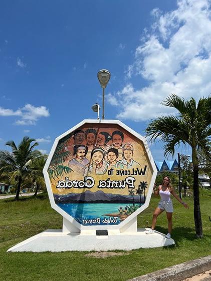 Ana Maria Castellanos poses in front of a sign welcoming visitors to Punta Gorda